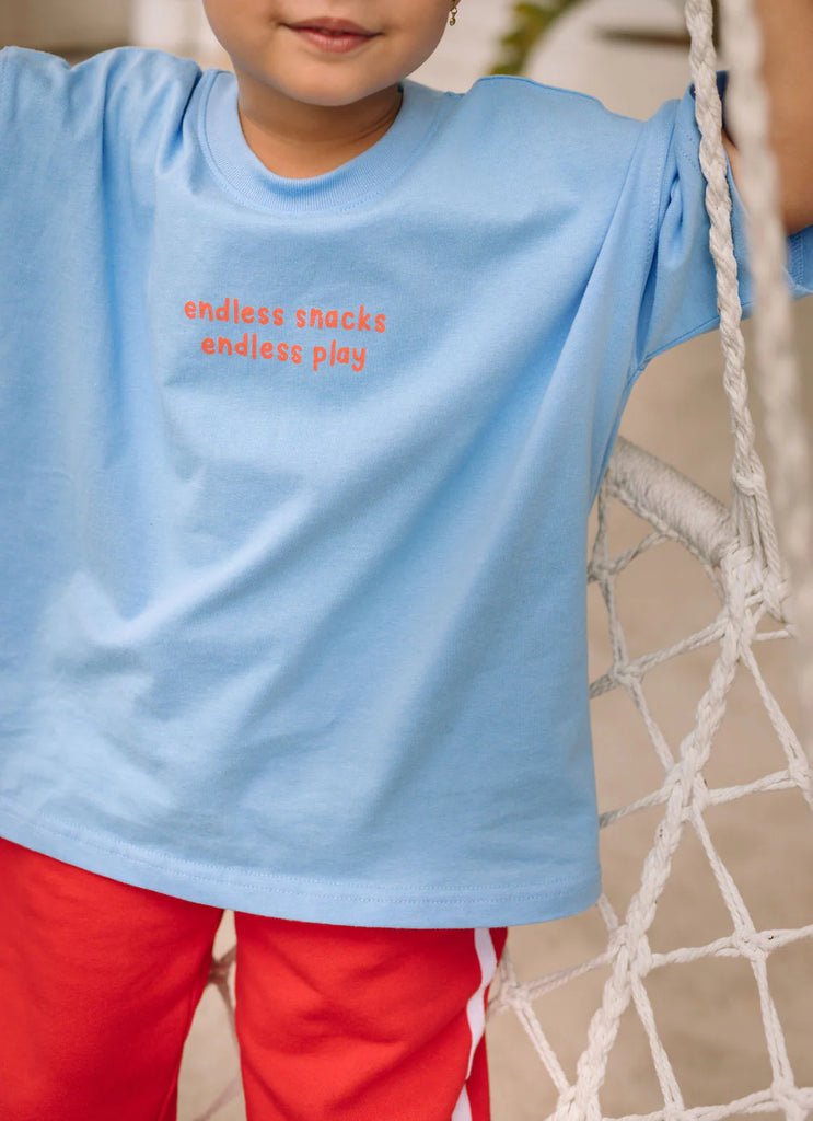 Child wearing a light blue t-shirt with text "endless snacks endless play", standing on a swing.