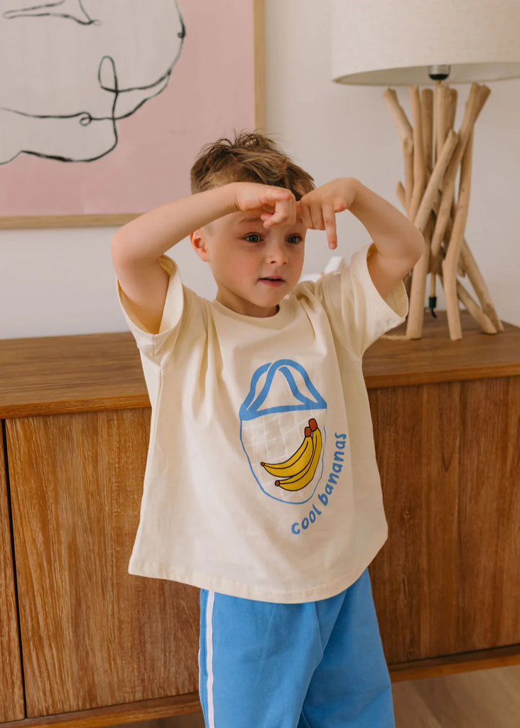 Child wearing a t-shirt with a banana graphic and text saying "cool bananas", standing in a room with wooden furniture and a lamp.